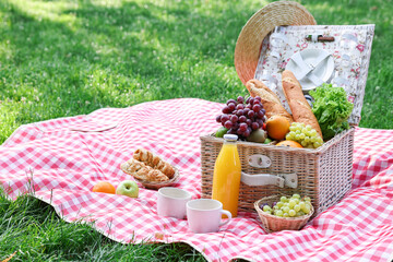 Picnic wicker basket with delicious food, drink and tableware on checkered blanket outdoors