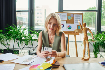 A blonde woman in a green sleeveless shirt is sitting at a desk in an office, checking her phone...
