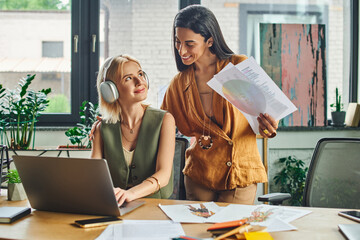 Two women in smart casual attire collaborate in modern office - one reviews documents, the other is focused on laptop