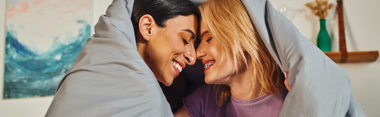 A happy lesbian couple cuddles under a blanket in their home, sharing a sweet moment.