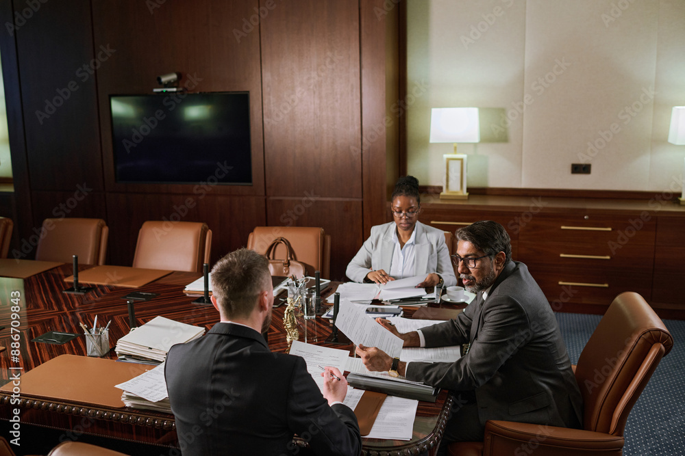 Poster group of intercultural coworking lawyers in formalwear sitting by large table in boardroom and worki