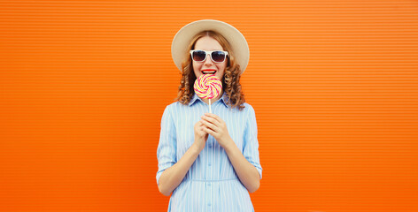 Summer portrait of happy young woman with colorful lollipop, cheerful girl in tourist hat on orange