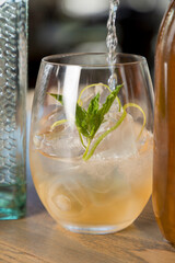 Gin being poured into a gin cocktail in a low ball tumbler with ice and a mint leaf.