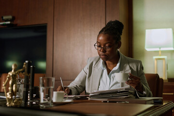 Young serious female notary with cup of coffee in hand sitting by table in boardroom and looking...