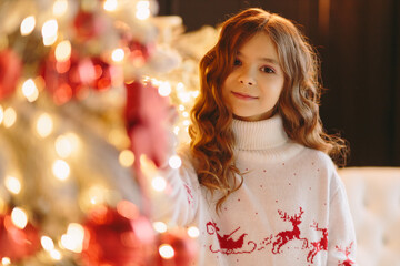 A cute girl in a white sweater with deer looks at the camera against the background of a Christmas tree.