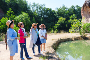 Group of elderly women exploring nature, wearing hats and casual clothing. Standing beside a pond in a rural area, enjoying the sunny day. Smiling, talking, and holding water bottles.