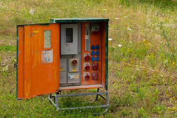 An old orange electrical box with a metal door open, revealing a variety of electrical outlets, switches, and a meter. The box is located in a field with green grass and yellow wildflowers.