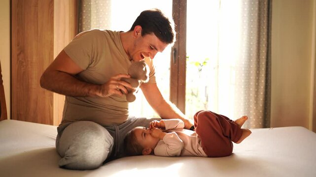 Dad plays with a laughing little girl lying on the bed with a teddy bear. High quality 4k footage