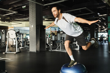 A muscular man creatively performs exercises on an exercise ball in a gym.