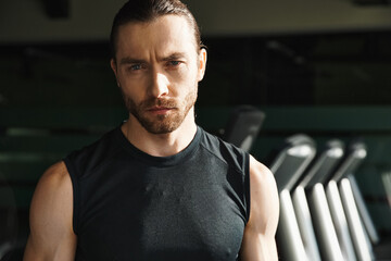 Obraz premium An athletic man in a black shirt stands confidently in front of a row of treadmills in a gym.
