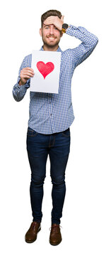 Young handsome man holding paper with red heart stressed with hand on head, shocked with shame and surprise face, angry and frustrated. Fear and upset for mistake.
