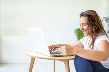 woman in casual wear working online, sitting on the floor and using laptop against white studio wall