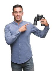 Handsome young man looking through binoculars very happy pointing with hand and finger