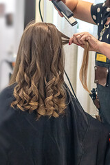 Naklejka premium Hair stylist prepares woman makes curls hairstyle with curling iron. Long light brown natural hair