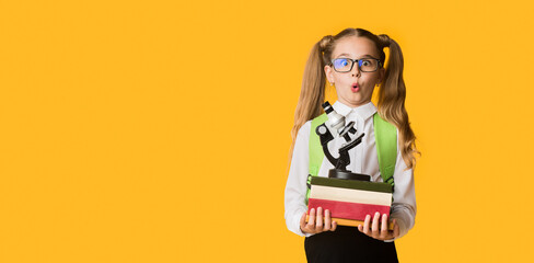 Back To School. Surprised First-Grade Schoolgirl Holding Stack Of Heavy Books And Microscope On...