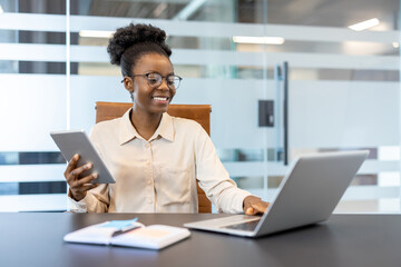 Smiling businesswoman using digital tablet and laptop in modern office environment. Confident...