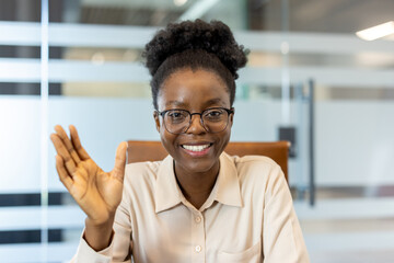 Smiling woman waving during video call in modern office. Friendly professional engaging in remote communication. Positive connection, casual yet professional ambiance, indicating ease and