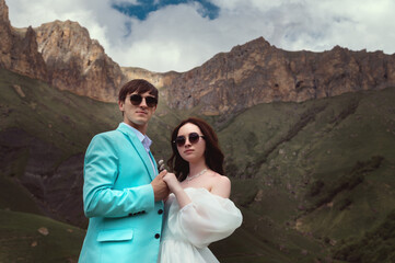 wedding couple in sunglasses and wedding suits stands stylishly against the background of high rocky mountains covered with grass.