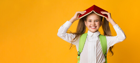 Home School Reading. Positive Elementary Schoolgirl Holding Book On Head Smiling To Camera Over Yellow Background. Studio Shot