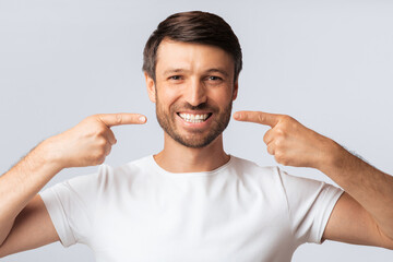 A man with a beard smiles widely and points to his teeth with both index fingers. He is wearing a...
