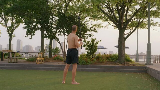 young athletic man in shorts do Steel Mace Exercises in public garden during sunset view . Lifestyle in New York. Sport workout  movement in Hudson river park Manhattan