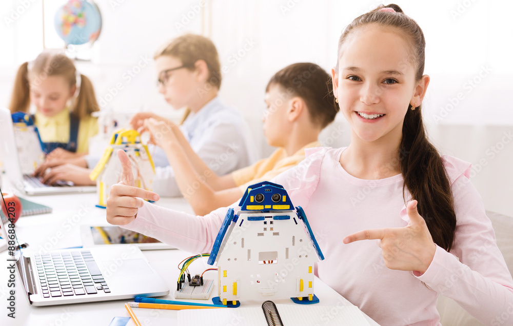 Poster A young girl smiles proudly as she points to a robot she has built in a classroom setting. Other students are visible in the background, suggesting that she is part of a class or group project