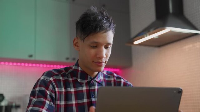 Biracial young man uses digital tablet computer while sitting at table in his stylish modern kitchen. Handsome young adult smiling guy working from home at digital device in apartment