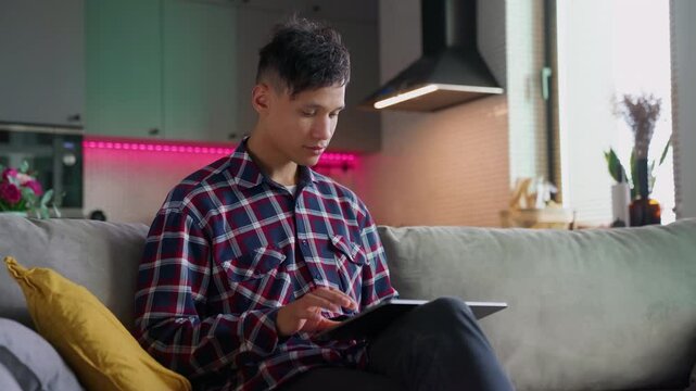 biracial young man use digital tablet computer sitting on sofa. Handsome smiling guy work at home