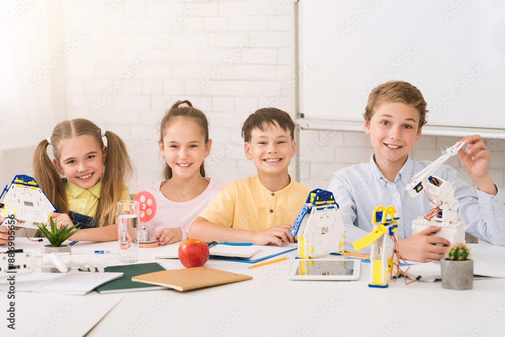 Fototapete Four young students are smiling and working on a robotics project in a classroom. They are surrounded by various robotic components, including a robotic arm and a robot.
