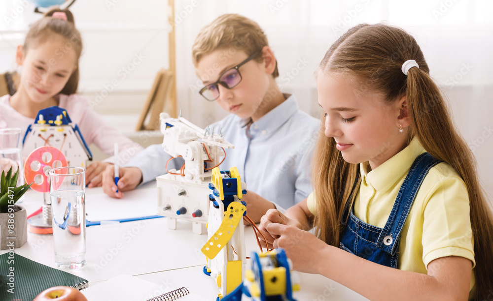 Aufkleber Three young students are sitting at a table in a classroom setting, working on building robots. They are concentrating on their work and seem to be enjoying the activity.
