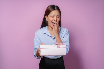 Young asian woman happily surprised holding a gift box against a pink background, radiating joy and excitement. Perfect for festive celebrations