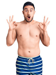 Fototapeta premium Young handsome man wearing swimwear looking surprised and shocked doing ok approval symbol with fingers. crazy expression