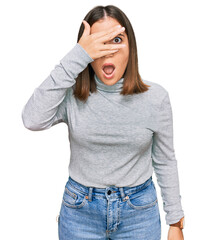 Fototapeta premium Young beautiful woman wearing casual turtleneck sweater peeking in shock covering face and eyes with hand, looking through fingers with embarrassed expression.