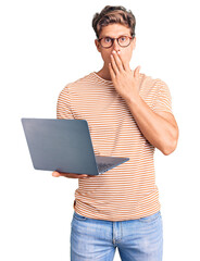 Young handsome man wearing glasses holding laptop covering mouth with hand, shocked and afraid for...