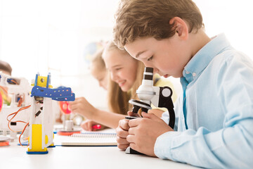 A young boy leans over a microscope in a classroom setting. He is intently looking through the lens while holding the microscope steady with both hands. His classmates are in the background