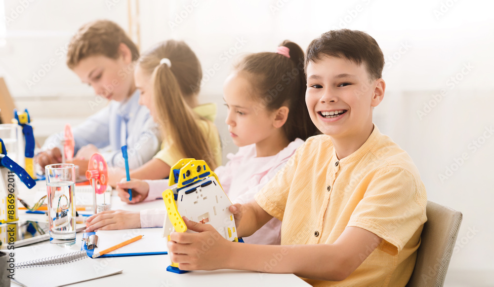 Aufkleber A young boy smiles while building a small robot during a STEM activity in his elementary school classroom. His classmates are working on other projects in the background.