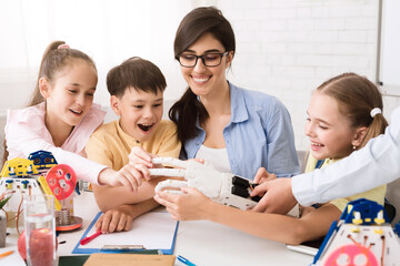 Three elementary school students are learning about robotics with their teacher. The teacher is holding a robotic arm and the students are interacting with it. They look excited and engaged
