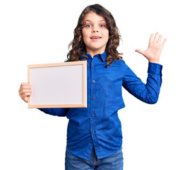 Cute hispanic child with long hair holding empty white chalkboard celebrating victory with happy smile and winner expression with raised hands