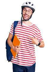 Young hispanic man wearing student backpack and bike helmet holding binder smiling happy pointing with hand and finger