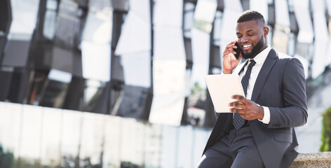 Positive African Entrepreneur Talking On Cellphone Using Digital Tablet During Coffee Break In City Urban Area