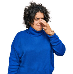 Young hispanic woman with curly hair wearing turtleneck sweater smelling something stinky and...