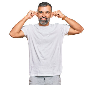Middle age handsome man wearing casual white tshirt smiling pulling ears with fingers, funny gesture. audition problem
