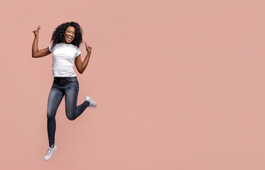 Black woman with dark skin and curly hair jumping in front of a solid pink background. She is wearing a white t-shirt and dark blue jeans. The woman is smiling and looks joyful.