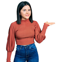 Young brunette woman with blue eyes wearing elegant sweater smiling cheerful presenting and pointing with palm of hand looking at the camera.