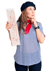 Young beautiful blonde woman wearing french beret holding city map serious face thinking about...