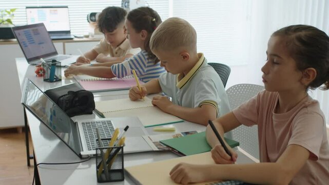 Medium shot of diverse schoolchildren sitting at desk when doing test during programming class using laptops at school