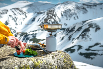 A woman's hands are cutting tomatoes to prepare food on a tourist gas stove during a mountain camping trip.