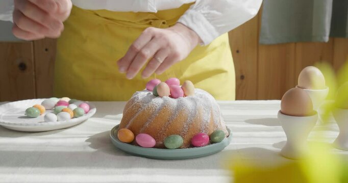 Female hands decorating Easter cake with small colorful chocolate eggs and daisy flowers. Anonymous woman making traditional easter cake or sweet bread with topping. Easter treat.
