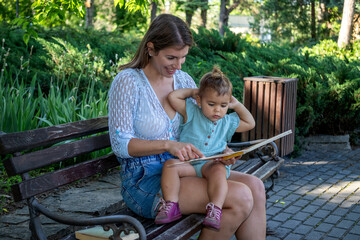The mother is sitting with her baby daughter on a bench in the park and reading a book together....
