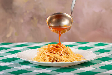 Pasta, tomato sauce being placed on a portion of spaghetti noodles, on a white plate, on a green...